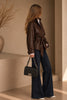 Chanel Boy Medium Black Lambskin — lifestyle photo: a woman in a belted brown leather jacket and white collared shirt over dark wide-leg jeans holding the black quilted lambskin Boy bag by its chain-and-leather strap, ruthenium CC turn-lock visible on the front flap.
